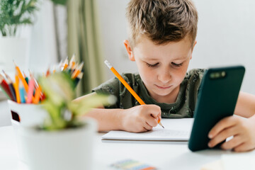 Wall Mural - Back to school. Distance learning online education. Caucasian smile kid boy studying at home with mobile smart phone and doing school homework. Thinking child with cellphone, pencil and training books