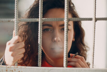 Young Brunette Curly Woman In Orange Suit Behind Jail Bars Smokes Cigarette. Female In Colorful Overalls Portrait