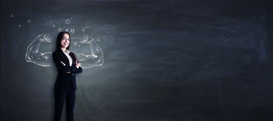 Attractive young european businesswoman standing on chalkboard wall background with drawn muscle hands and mockup place for youre advertisement. Strength, leadership and independence concept.