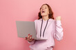 Portrait of enthusiastic curly haired teenage girl in hoodie screaming with joy and holding laptop, rejoicing victory. Indoor studio shot isolated on pink background
