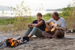 © ulza - Senior couple Camping holiday in the summer nature. Camping concept. senior man playing guitar and singing song to his wife sitting on the beach near the fire