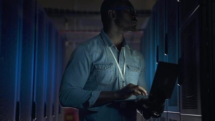 Young man working with laptop and typing while standing in dark server room spbas. Close-up view of African American male programmer holds device in hand and looks at screen, examines equipment and