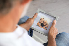 Senior Man Holding Frame With Photo Of His Wife And Granddaughter At Home, Closeup