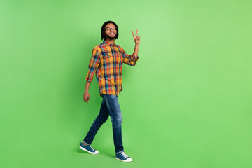 Wall Mural - Photo of pretty sweet dark skin guy dressed checkered shirt glasses walking showing v-sign empty space isolated green color background