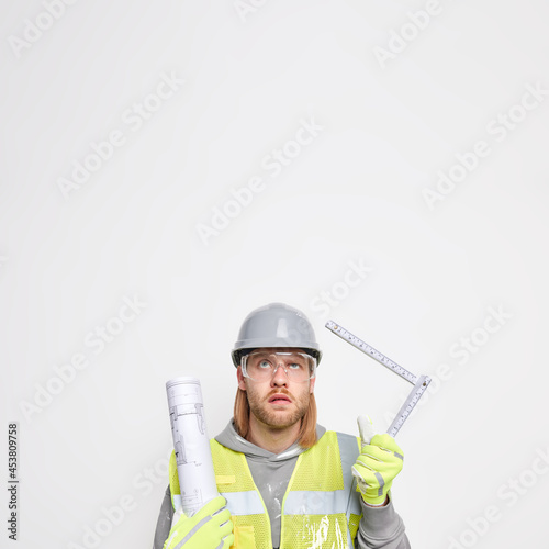 Vertical shot of ginger man maintenance worker holds tape measure and paper blueprint wears protective helmet uniform isolated over white background blank copy space. Industry and remodeling