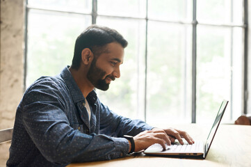 positive smart indian freelancer guy working on laptop at home office, websurfing, multiracial easte