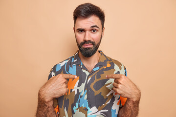 Wall Mural - Serious bearded man points at himself asks who me has attentie look at camera wears coloful shirt surprised being chosen isolated over bege background. Handsome adult guy asks about himself.