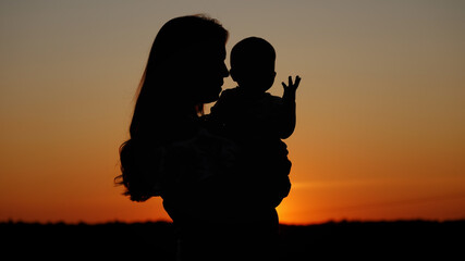 Caring mother holds the baby in her arms and kisses him, unrecognizable silhouette at sunset, motherly love