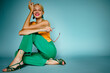 Happy smiling fashionable woman wearing trendy green color wide leg jeans, strappy sandals, posing, sitting on blue background. Copy, empty space for text