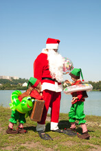 Santa Claus On Vacation Near The Water In The Summer With The Kids Elves. Children Have Inflatable Circles And Cute Smiles. The Concept Of Rest In Warm Countries