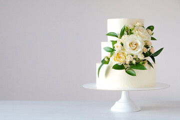 Wall Mural - Three-tiered white wedding cake decorated with flowers and green leaves on a white wooden background.