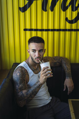 Sticker - Vertical shot of a young caucasian guy with tattoos sitting in the cafe and drinking a cup of juice