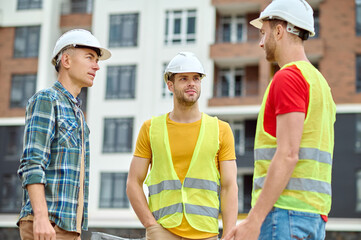 Wall Mural - Three builders in protective helmets having a conversation outdoors