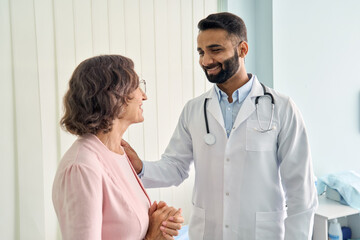 Wall Mural - Happy young Indian doctor therapist in white coat has appointment consulting supporting putting hand on shoulder of older senior female patient in modern clinic hospital. Medical healthcare concept.