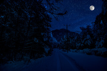 Mountain Road through the snowy forest on a full moon night. Scenic night winter landscape of dark blue sky with moon and stars