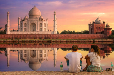 Fototapete - Taj Mahal Agra at sunset with moody sky and water reflection enjoyed by tourist couple sitting at Mehtab Bagh beside river Yamuna	