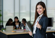 © Bangkok Click Studio - Portrait of young attractive Asian female office worker in formal business suits  smiling at camera in office with blurry colleagues sitting in office as background