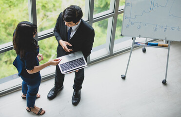 Top view flat lay shot of male and female professional Asian business worker standing at the windows and talking about a new company project with a whiteboard in the back. Businessman holding laptop