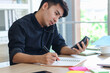 © Stella - Busy businessman in black shirt working on office desk, taking notes while talking with customer on mobile phone, and the other hand holding a calculator, writing information data on paper.