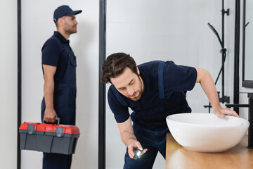 Wall Mural - plumber with flashlight checking sink in bathroom near blurred colleague with toolbox