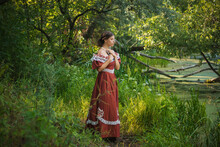 Sad Young Woman In A 19th Century Dress By The River With A Book In Her Hands. Summer Landscape. The Photo.