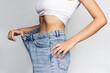 © Марина Демешко - Cropped shot of a young slender woman dressed in wide jeans and white top demonstrates successful weight loss isolated on a light color gray background. Diet and wellness concept