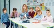 © VAKSMANV - Joyful Caucasian woman tutor sitting at classroom with little children constructing robot, looking at camera and smiling at science class. Technology lesson, studying, elementary school education