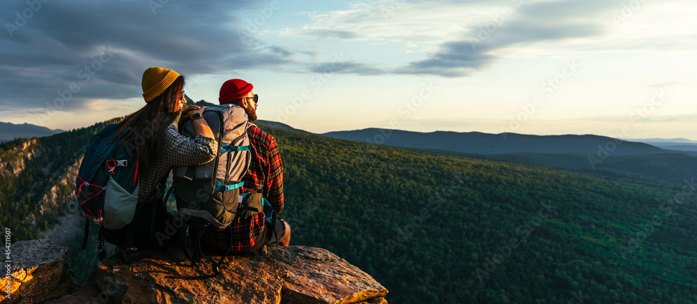 The couple meets the sunset in the mountains. Two travelers are sitting on the edge of a cliff and admiring the beautiful panoramic view. Travelers with backpacks at sunset in the mountains. Panorama - obrazy, fototapety, plakaty 