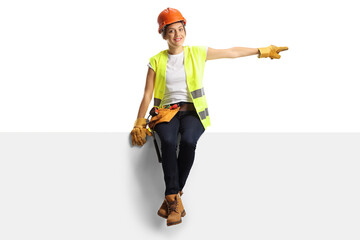 Wall Mural - Female construction worker wearing a hardhat and a tool belt sitting on a panel and pointing