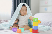 Asian Little Baby Boy Playing A Tower Cup Toy On A Bed In Bedroom. A Kid Playing And Smiling Under Covering A Blue Duvet. Child Development And Creativity Concept.