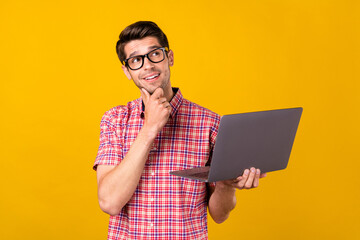 Photo of cheerful young happy man look empty space think hold laptop freelancer isolated on yellow color background
