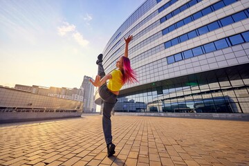 young woman in bright sports clothes and dyed pink hair has raised leg high and is training in city