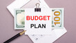 © Елена Дигилевич - On a gray background, a white calculator, a pen, banknotes and a sheet of paper under a black paper clip with the text BUDGET PLAN. Business concept