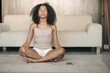 © Pictures by Alex - Young black woman practicing yoga at home, sitting in Easy Seat pose, her eyes closed, listens audio meditation by headphones with the phone