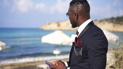 side view of confident happy african american handsome man in suit with boutonniere texting on smart
