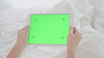 A girl with a beautiful manicure is lying in a snow-white bed and looking at the screen of a tablet with a green screen. A woman on self-isolation holds a tablet with a green screen for cg.