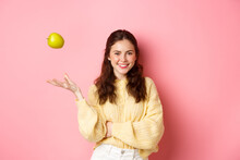 Young Smiling Woman Looking With Confidence, Throwing Apple In Air, Eating Healthy Fruits To Keep That Smile Perfect, Standing Against Pink Background