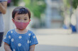 © TimmyTimTim - Asian boy toddler wearing red cloth face mask and blue shirt with white polkadot standing on the street outside of his house. Covid-19 (coronavirus) pandemic prevention during in child concept.