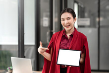 Front View Of Asian Businesswoman Wearing Red Dress Holding Pointing Tablet Blank White Screen. Looking At Camera. Mock Up.