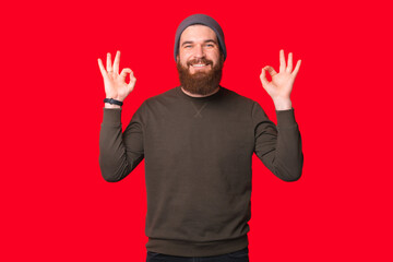 Wall Mural - Pleased bearded man is showing ok gesture over red background while smiling at the camera.