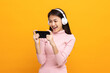 © Chanakon - Beautiful young asian women play mobile game and put on wireless headphone standing on isolated yellow background. Playing game on smartphone winning victory moment. Very enjoy and fun relax time
