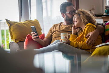 Tenderness time moment for mature caucasian couple at home sitting on the couch together - love and relationship adult people indoor lifestyle - man kissing woman with phone on hand