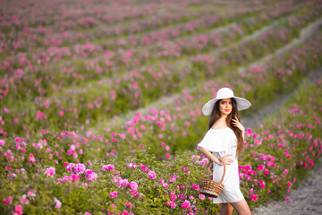 Wall Mural - Beautiful young woman portrait in white hat over roses field. Carefree happy brunette with healthy wavy hair having fun outdoor in nature.
