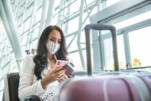 Woman in face mask checks her phone while waiting for an airplane in a hall, holding passport and a boarding pass
