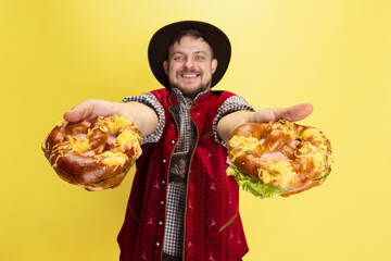 Wall Mural - Happy smiling man dressed in traditional Bavarian costume with yummy pretzels isolated over yellow background. National cuisine, holidays, traditions