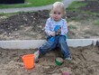  a sad child against the background of a poor, unfurnished playground. offended child in the sandbox, with a scoop, mold and bucket