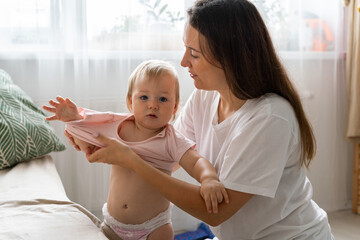 Adorable blond blue-eyed curious baby looking at camera while being dressed up by nanny in casual clothing at home near sofa. Concept of child care. Horizontal shot.