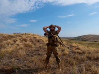 Photo of a mercenary sniper in camouflage clothes under the scorching sun. He stands with a rifle and surveys the area.