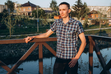 A young man in a plaid shirt at a street photo shoot at sunset, near a pond. 