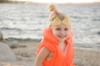 © triocean - Toddler girl in orange life jacket vest with water behind
